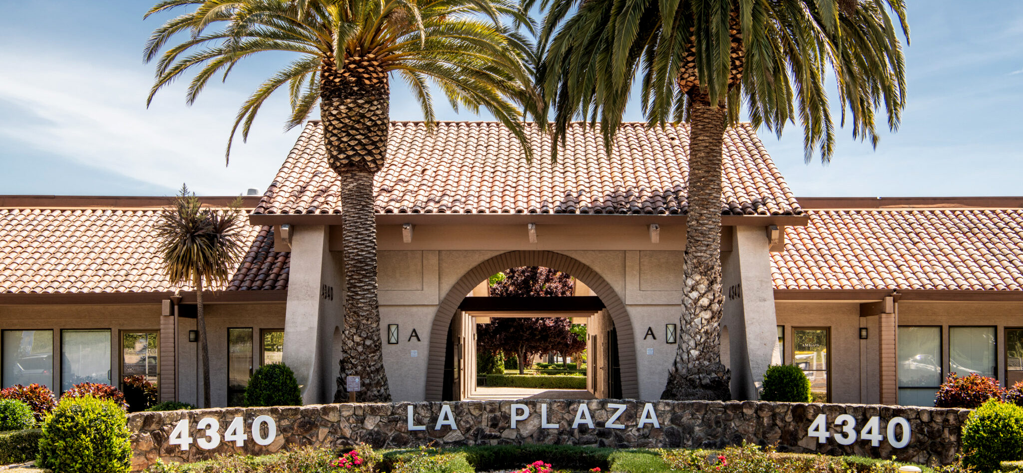 Exterior view of La Plaza building with tiled roof, stone signage, and palm trees at the entrance, located at 4340.